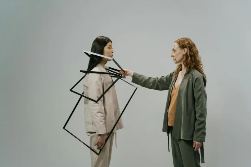 Young female model in green costume and beige top putting empty photo frames on shoulders of other female model wearing light beige jacket and trousers
