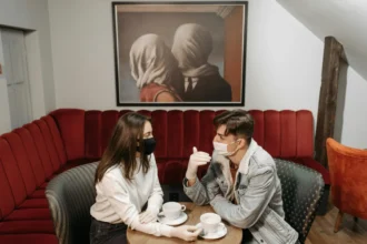 A couple enjoys coffee while wearing masks in a cozy café, highlighting the pandemic's impact.