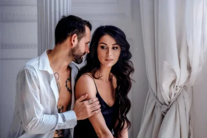 Calm young boyfriend in unbuttoned shirt hugging attractive woman wearing black silk nightgown while spending sunny morning together in light room