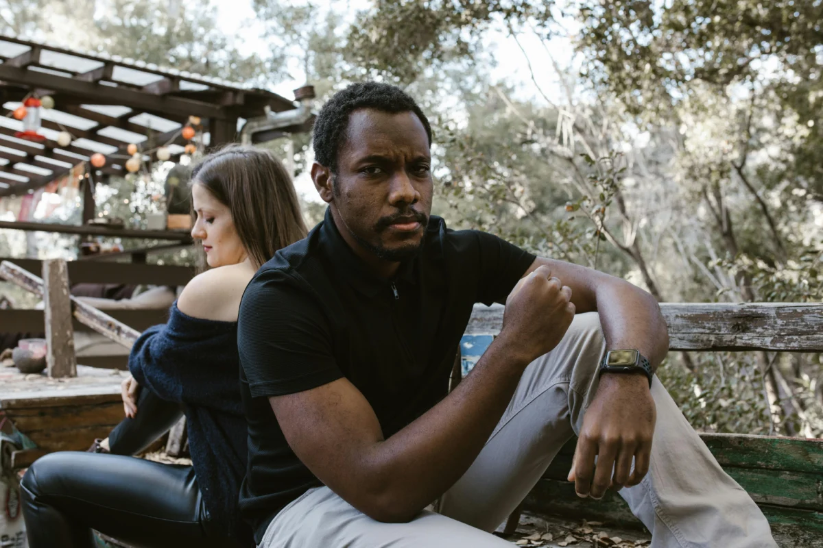 A thoughtful man sits outdoors, with a woman sitting distantly behind him.