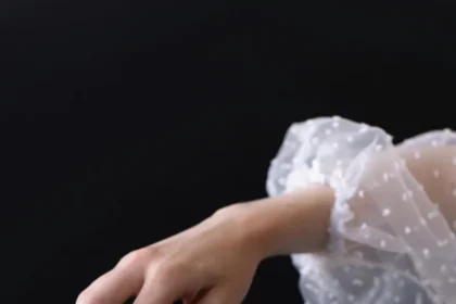 Delicate hand in white sleeve holding a red apple against a dark background, symbolizing temptation and elegance.