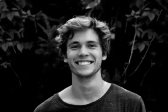 A black and white portrait of a young man smiling outdoors with natural background.