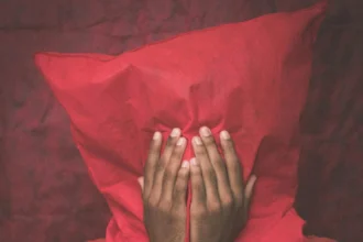 Moody portrait of an adult hiding behind a red pillow with hands covering their face.