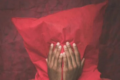 Moody portrait of an adult hiding behind a red pillow with hands covering their face.