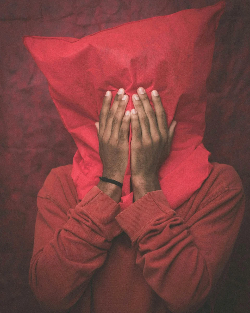 Moody portrait of an adult hiding behind a red pillow with hands covering their face.