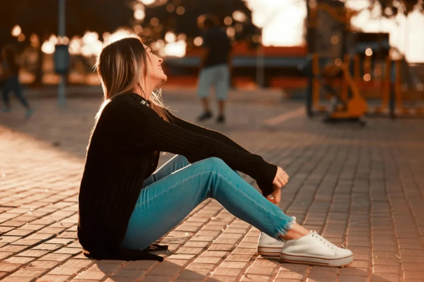 A young woman sitting on the ground outdoors, enjoying a sunny day with a smile.