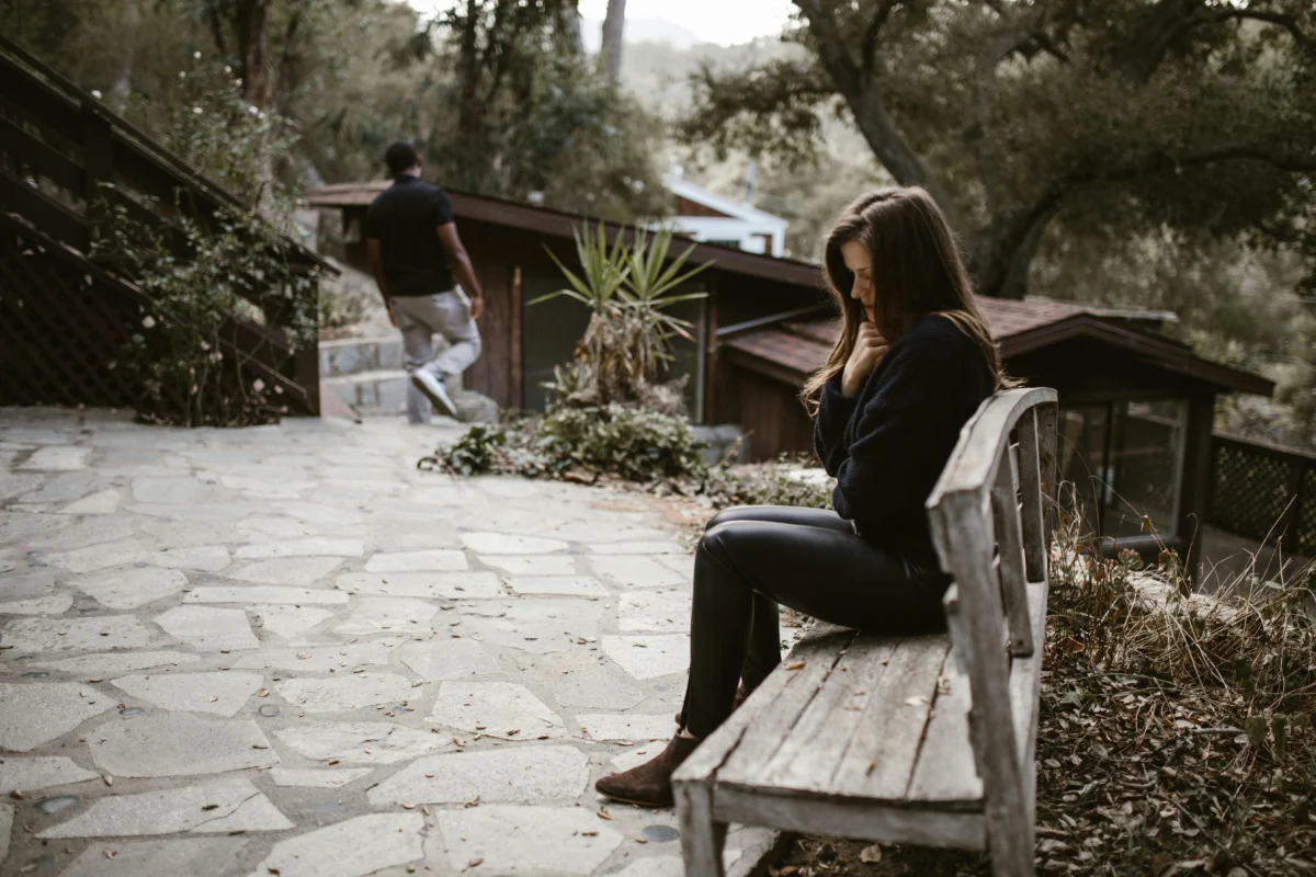 A woman sits pensively on a bench outside after a breakup, while a man walks away.