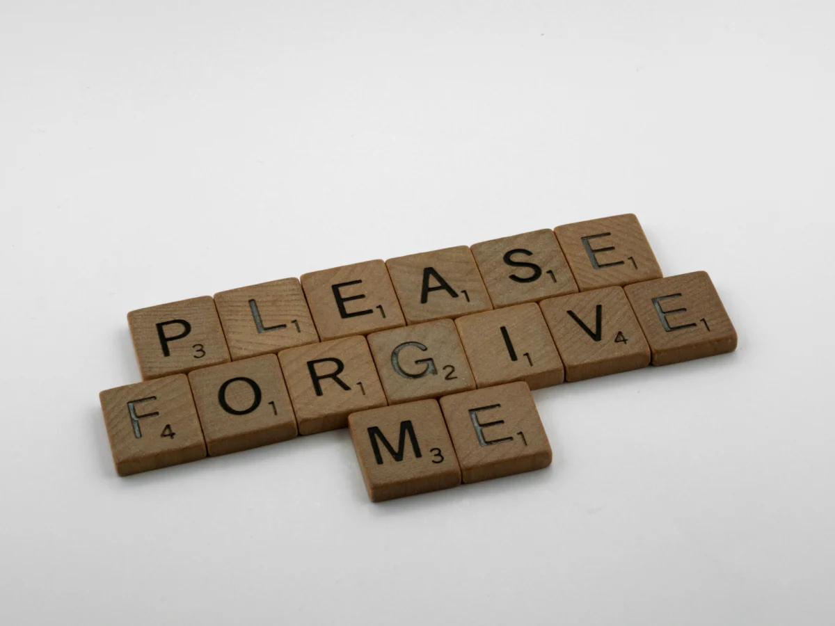 Wooden Scrabble tiles spelling 'Please Forgive Me' on a white surface, emphasizing forgiveness.